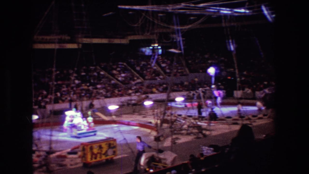 1974 Boston 8mm frame showing an indoor circus ring from the audience area, with spectators, rigging, spotlights, and a performer near a decorated cart.