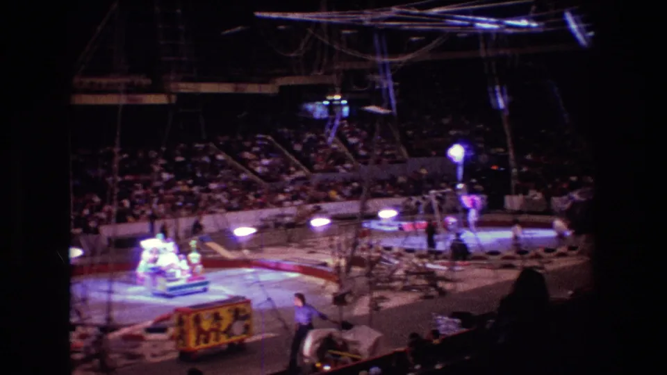 1974 Boston 8mm frame showing an indoor circus ring from the audience area, with spectators, rigging, spotlights, and a performer near a decorated cart.