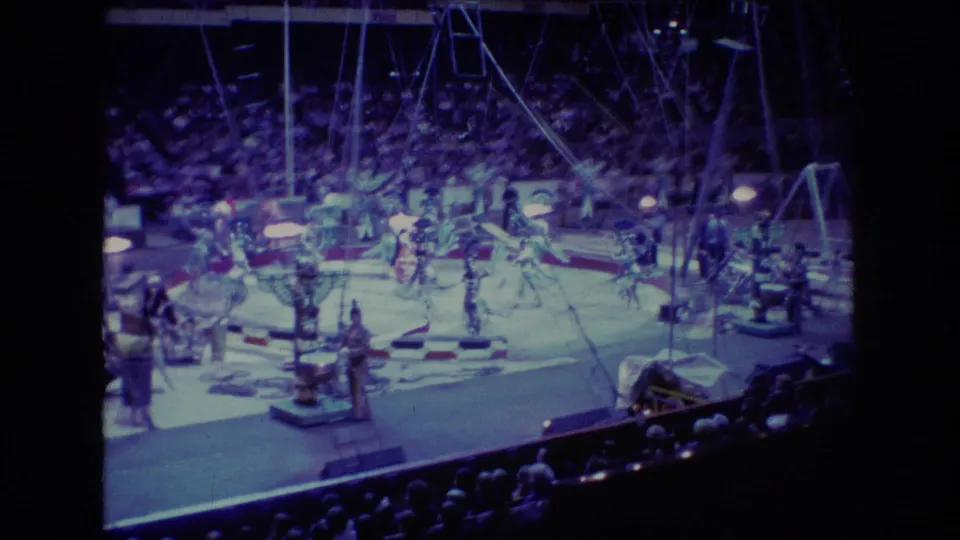 Wide 1974 Boston home-movie view of a circus performance with several costumed figures, arena seating, blue-tinted lighting, ladders, and overhead rigging.