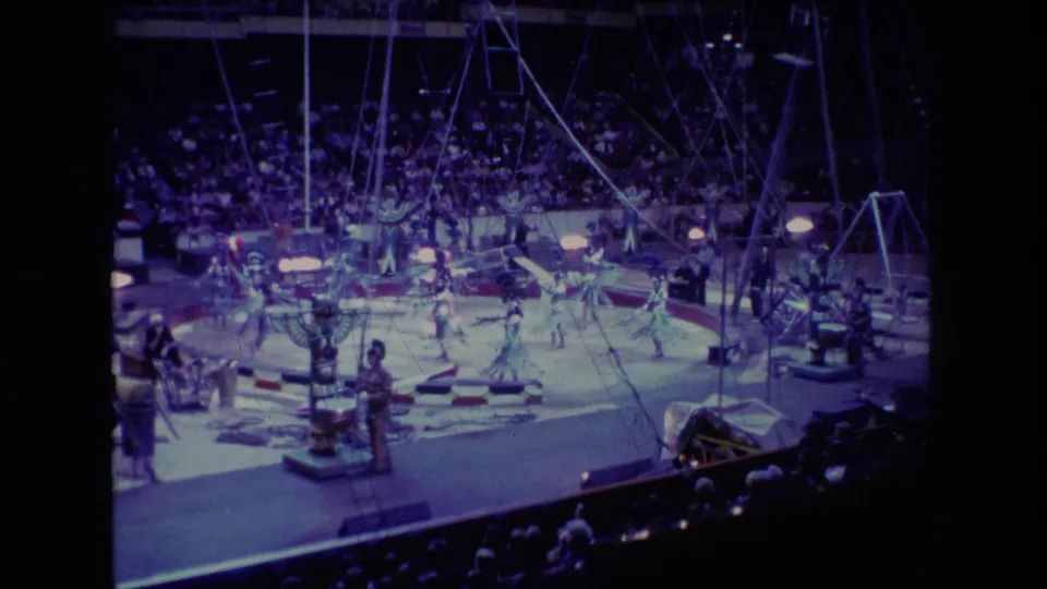 1974 Boston 8mm frame of an indoor circus act, with performers across the ring, spectators in the background, rigging above, and stage equipment around the floor.