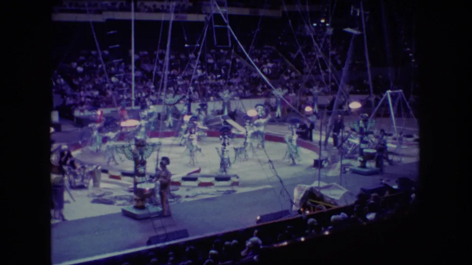 1974 Boston 8mm home-movie frame showing a wide view of circus dancers and acrobats performing under rigging before a large arena audience.