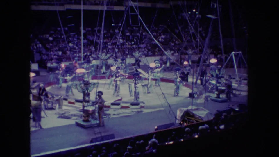 1974 Boston circus footage frame with dancers and acrobats spread across the arena floor while spectators watch from tiered seating.