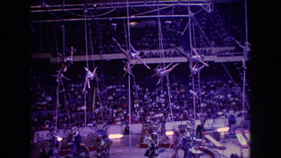 1974 Boston circus frame showing aerial performers suspended on ropes above the ring, with rigging, stage lights, and a large audience in the background.