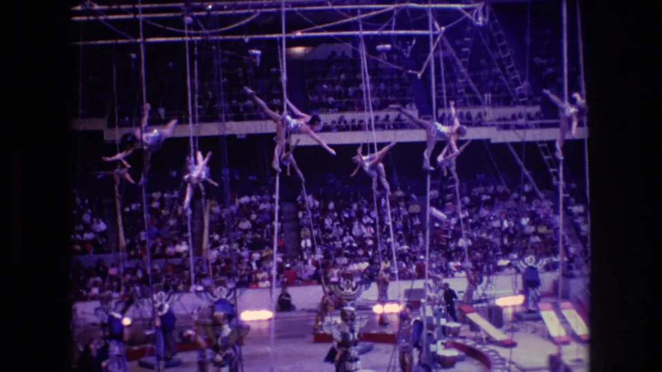 In 1974 Boston 8mm footage, several circus aerialists hang from ropes beneath overhead rigging while spectators fill the arena seating behind them.