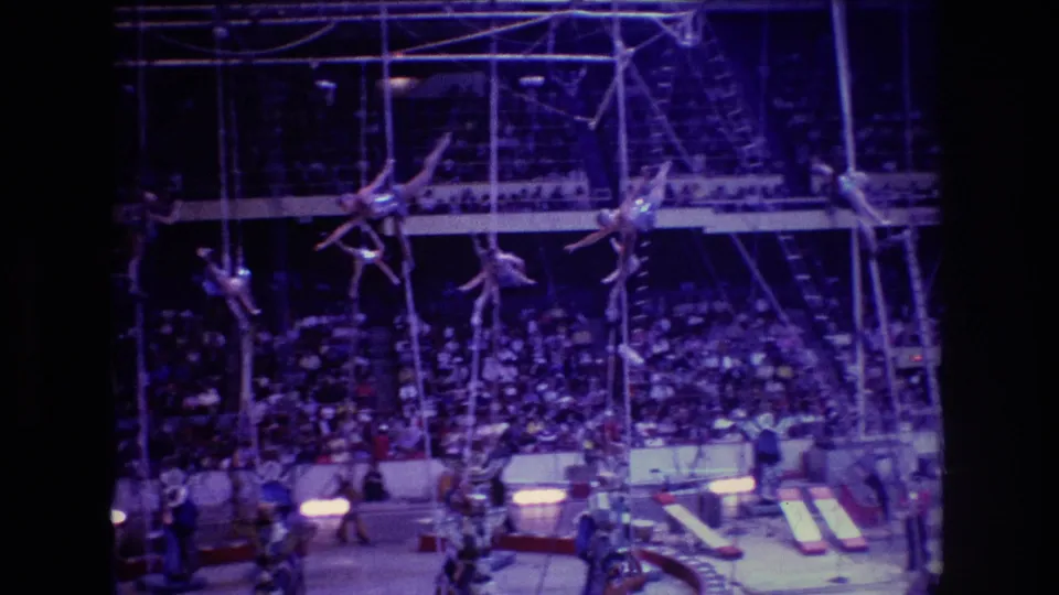 The 8mm frame shows multiple aerialists spread across the rigging, with performers or crew below and rows of spectators watching from the arena seats.