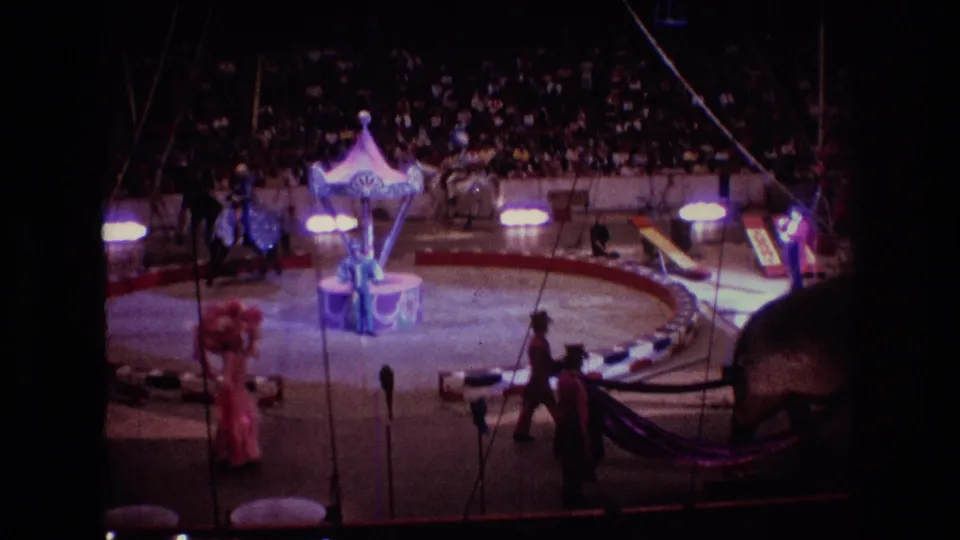 1974 Boston home-movie frame of a circus ring under bright lights, with performers circling near a central prop, audience members in the stands, and the elephant partly visible at the edge.