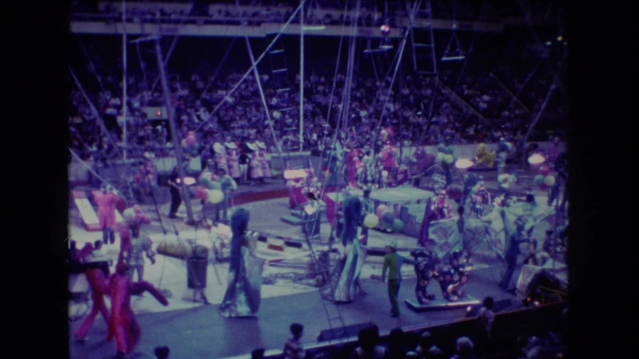 Costumed circus performers move around a ring while a large audience watches from the stands.