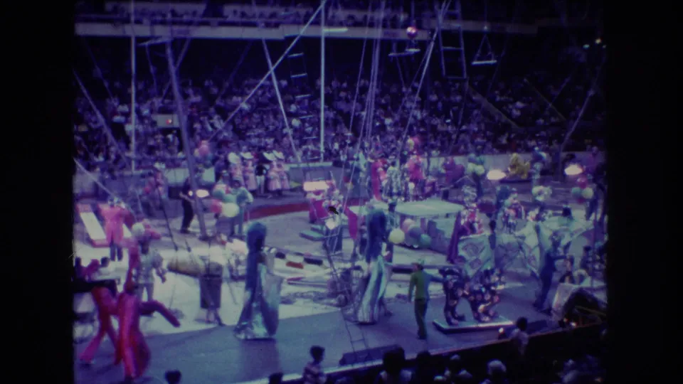 1974 Boston 8mm frame showing costumed circus performers, clowns or clown-like figures, props, balloons, rigging, and a stadium audience under purple lighting.