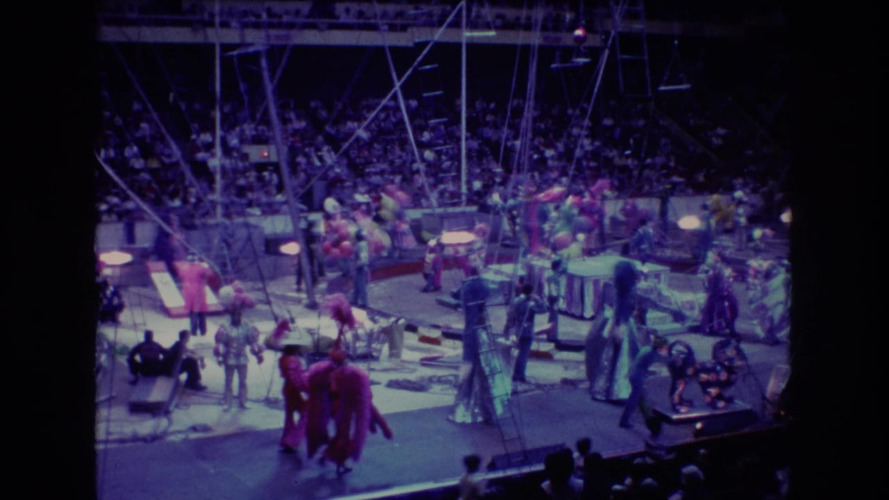 Performers in bright costumes parade through the circus ring with audience seating behind them.