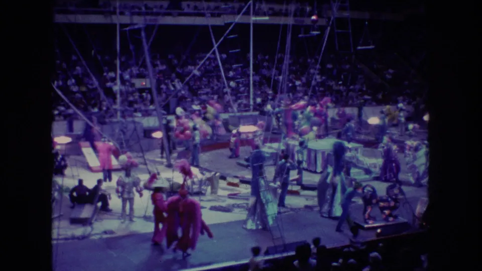 1974 Boston 8mm frame of a circus parade in an arena, with red-costumed figures, props, balloons, overhead rigging, and spectators in the background.