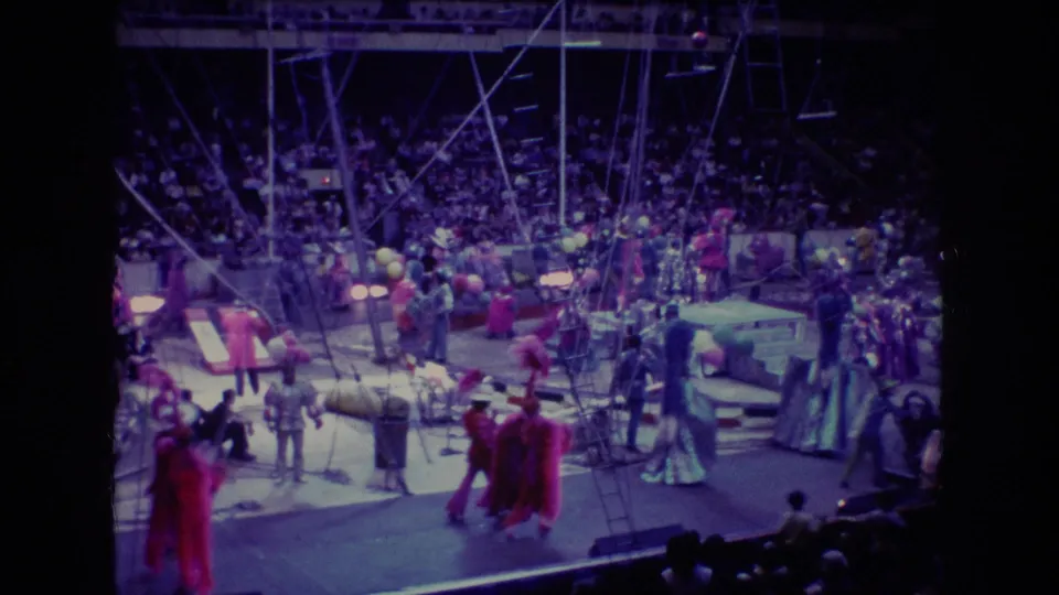1974 Boston Super 8 frame showing a busy circus ring with multiple costumed performers, clown-like figures, stage props, balloons, rigging, and packed stands.