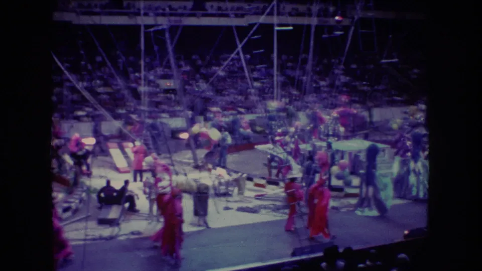1974 Boston 8mm frame with motion-blurred circus performers moving across an arena ring, surrounded by props, rigging, balloons, and spectators.