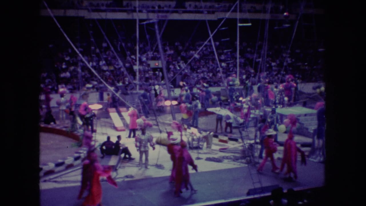 Circus performers in red costumes move through a dimly lit main ring with rigging and audience seating behind them.