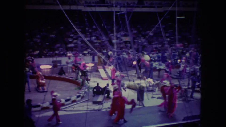 1974 Boston home-movie frame shows performers in red and pink costumes near circus equipment in the main ring, with audience members seated in the dim arena behind them.