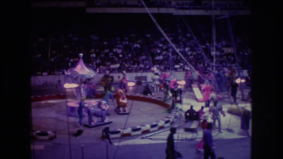 1974 Boston home-movie footage shows performers moving across the circus ring near set pieces and aerial rigging, with a large seated audience in the background.