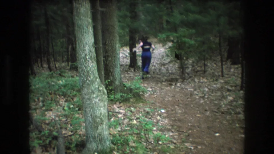 A person in blue pants walks away along the wooded Baxter, Maine trail while trees, undergrowth, and a dirt path fill the surrounding frame.