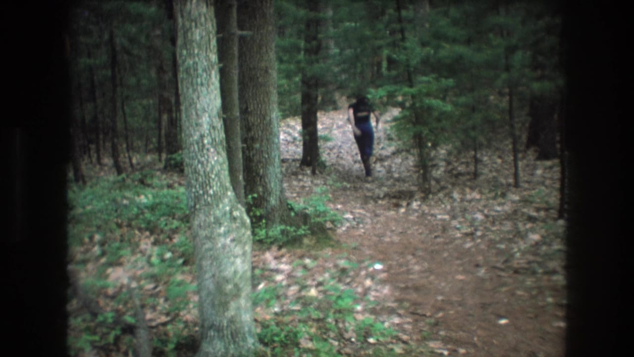 1975 Baxter, Maine 8mm frame showing a distant person walking away along a dirt trail through dense woodland, bordered by dark viewfinder edges.