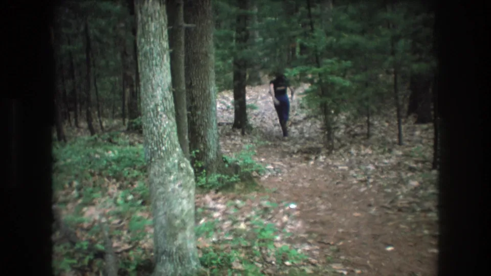 1975 Baxter, Maine 8mm frame showing a distant person walking away along a dirt trail through dense woodland, bordered by dark viewfinder edges.