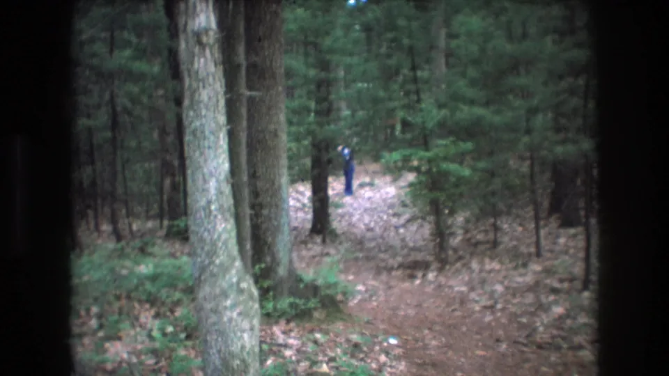 1975 Baxter, Maine 8mm frame showing a distant person in blue on a brown dirt path surrounded by tall trunks, fallen leaves, and green forest growth.