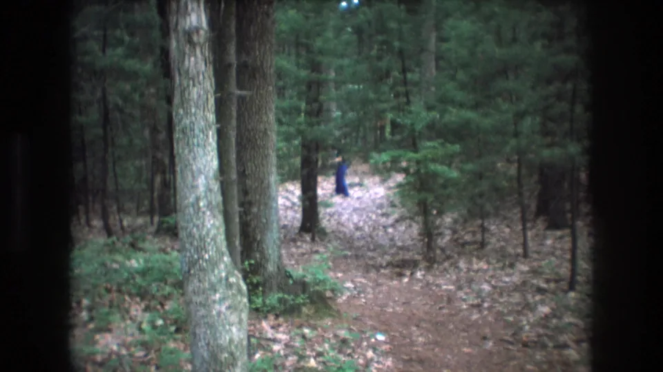 1975 Baxter, Maine 8mm frame with a distant blue-clothed figure on a brown forest path, framed by dark borders and surrounding trees.