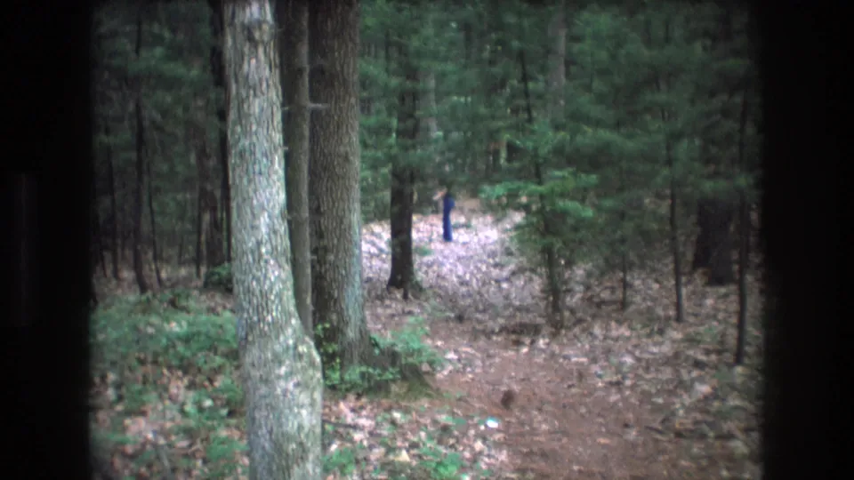 1975 Baxter, Maine 8mm frame showing a leaf-covered forest path, tall tree trunks, and a distant person in dark blue partly framed by dark vertical edges.