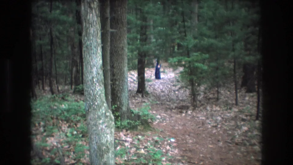 1975 Baxter, Maine home-movie frame with a distant person in dark blue moving along a forest trail, seen through dark vertical obstructions at the frame edges.