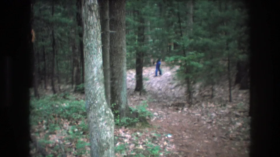 Soft, grainy 1975 Baxter, Maine frame showing a person in dark blue on a wooded trail, partly obscured by trees and dark frame borders.