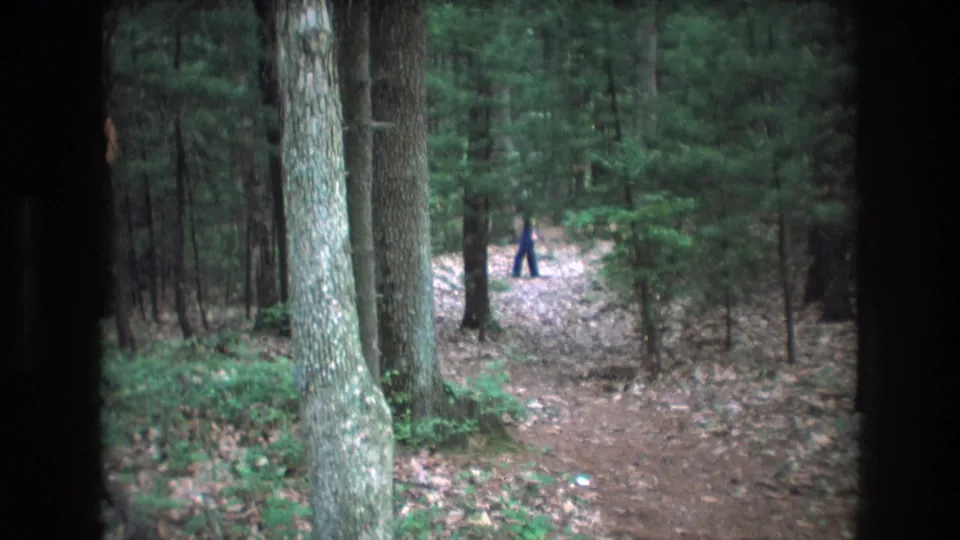 1975 Baxter, Maine 8mm frame showing a distant person in dark blue farther down a leaf-strewn forest trail, with dark vertical framing and dense trees around the path.