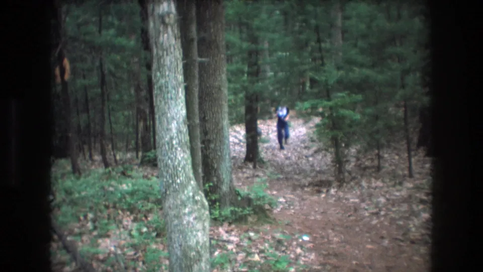 1975 Baxter, Maine 8mm frame showing a person in a light shirt and dark pants running along a wooded dirt trail toward the camera.