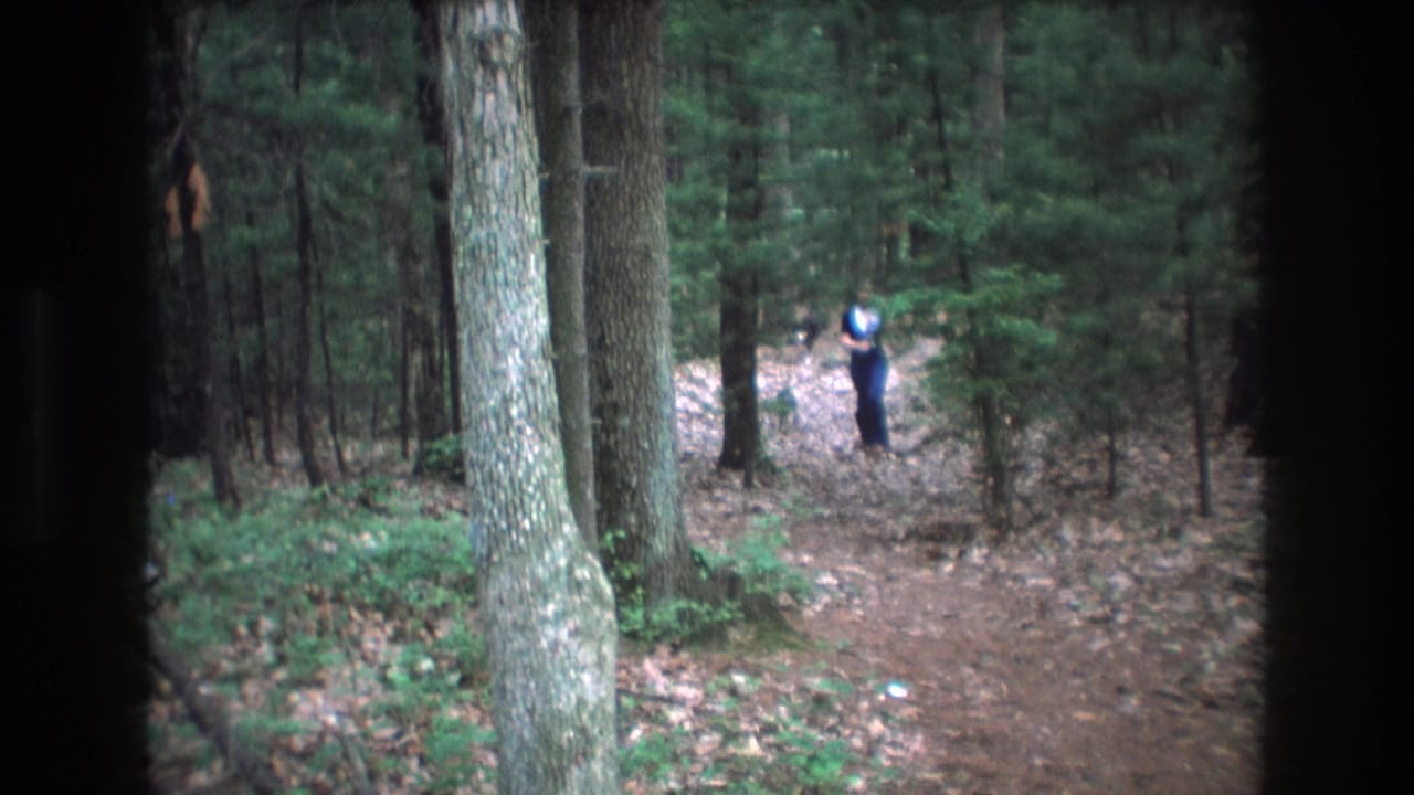 A distant young woman appears on a wooded trail in Baxter, Maine home-movie footage from 1975, framed by tall trees, leaf litter, and dark 8mm edge vignetting.