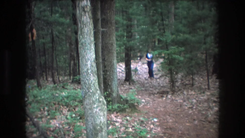 A distant young woman appears on a wooded trail in Baxter, Maine home-movie footage from 1975, framed by tall trees, leaf litter, and dark 8mm edge vignetting.