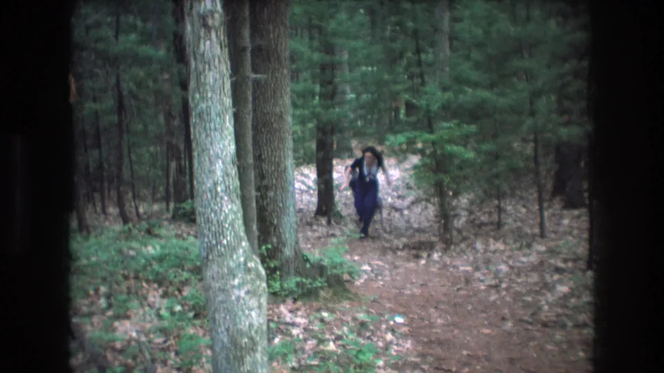 The young woman runs toward the camera along the Baxter, Maine forest path, with a small dog visible near the trail and dense trees behind her.
