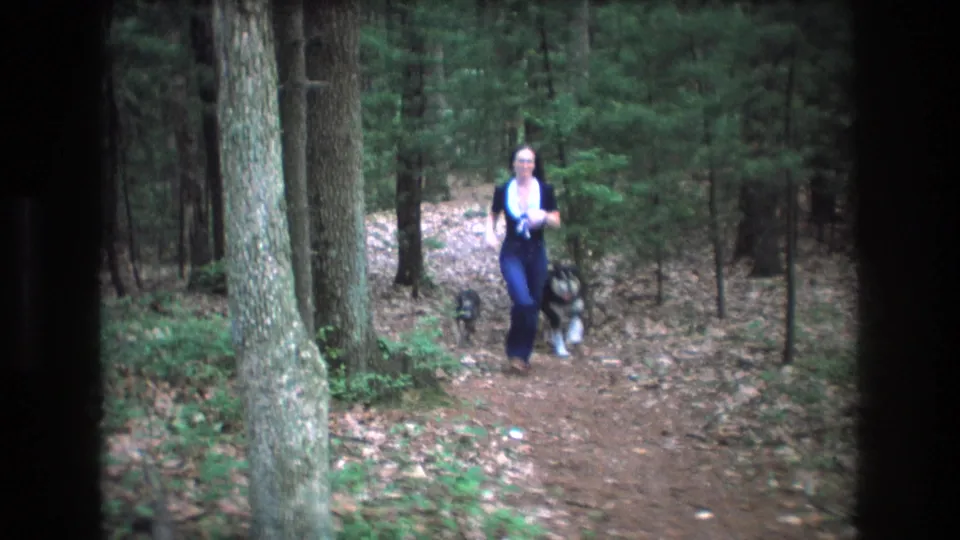 A young woman in dark blue clothing with a white collar runs toward the camera on a wooded Baxter, Maine trail, with two small dogs moving near her legs.