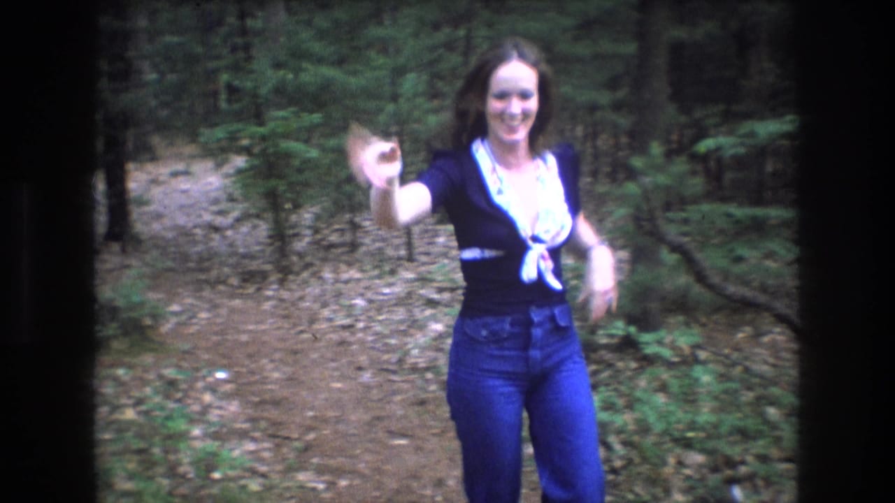 In metadata-supported 1975 Baxter, Maine home-movie footage, a woman in jeans waves toward the camera while standing on a leaf-strewn forest trail.
