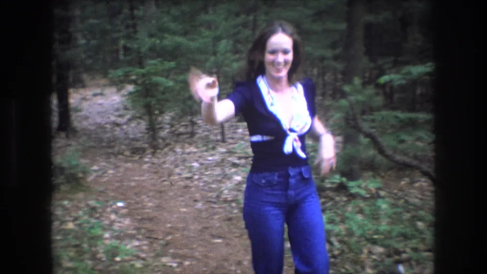 In metadata-supported 1975 Baxter, Maine home-movie footage, a woman in jeans waves toward the camera while standing on a leaf-strewn forest trail.