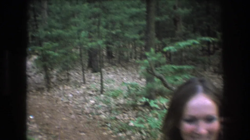 The woman moves close to the camera at the right edge of the 8mm frame, with the surrounding forest trail and trees still visible behind her.
