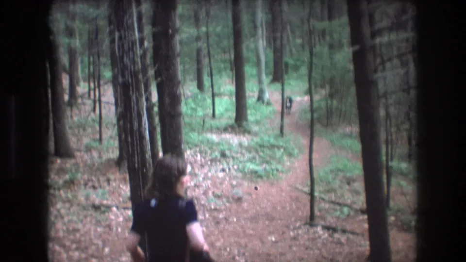 In metadata-supported Baxter, Maine in 1975, a woman in a dark blouse and blue pants runs along a forest trail carrying a dark handbag.