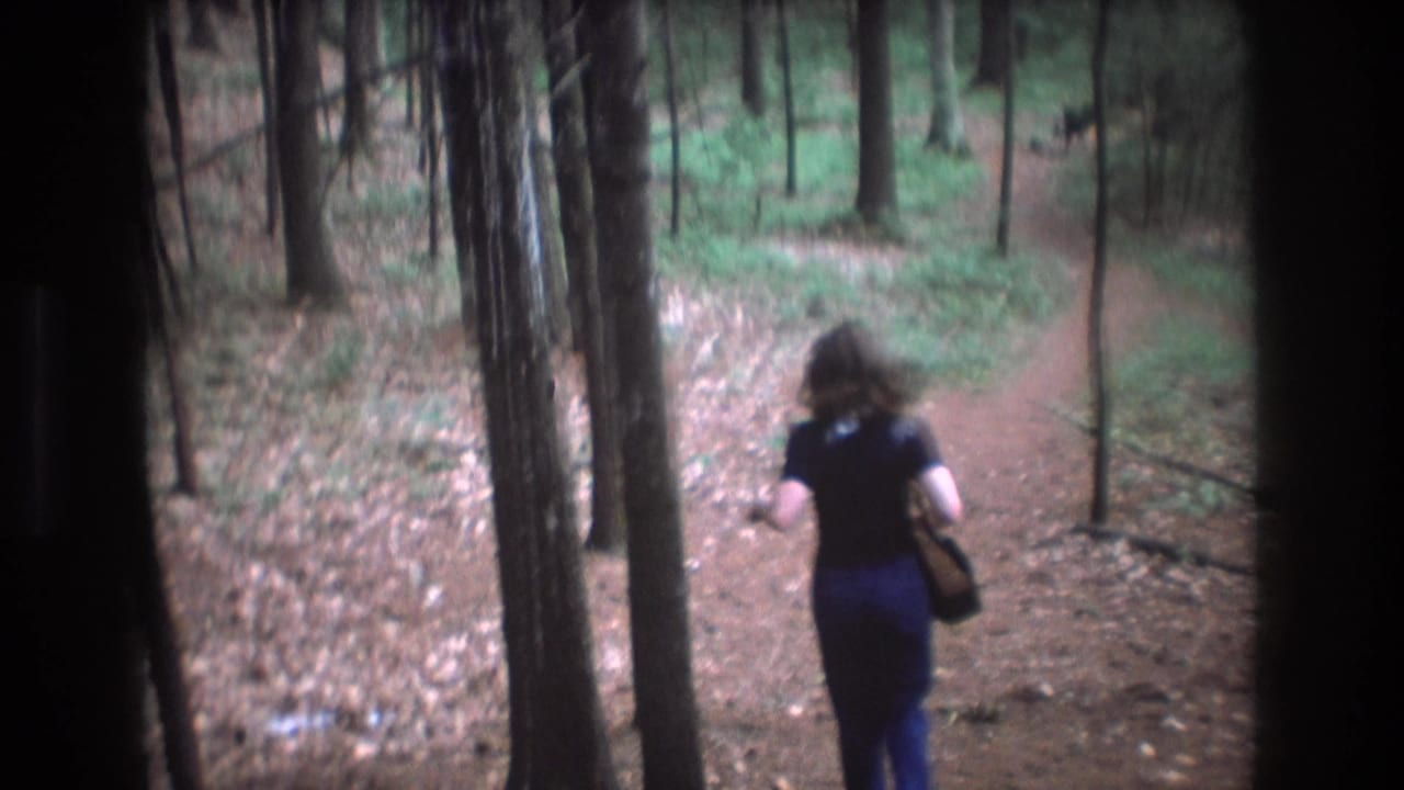 Woman runs away down a dirt forest path between tall trees while carrying a dark handbag.