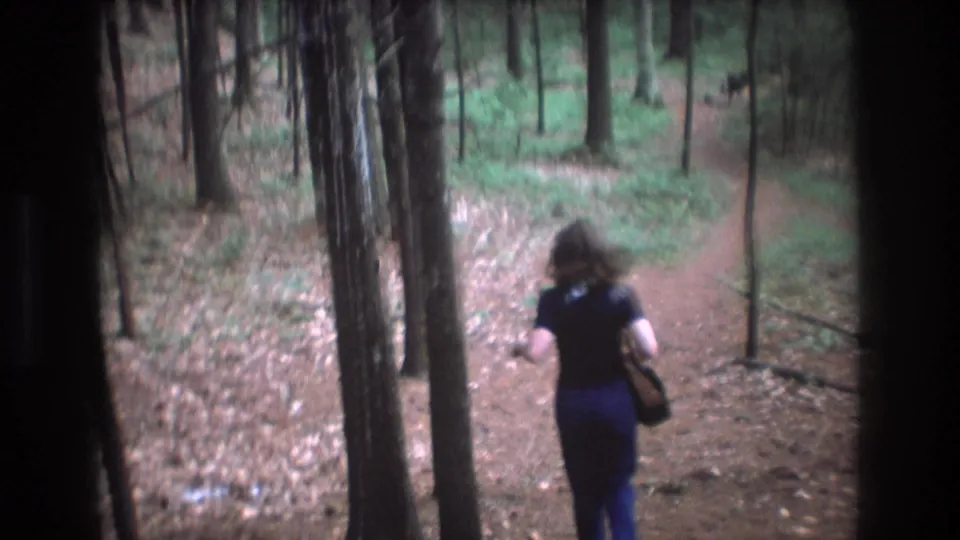 The woman continues down the Baxter, Maine forest path, seen from behind in soft 8mm color as the trail curves through trees.