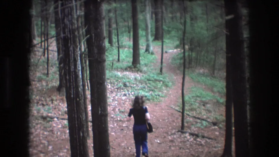 A woman runs along the dirt trail in the 1975 Baxter, Maine home-movie footage while a small dog appears behind her on the forest path.