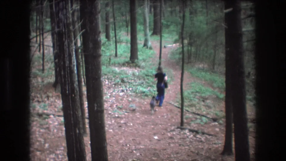 1975 Baxter, Maine 8mm frame showing a woman in a dark top and blue pants walking a small dog along a forest trail between tall trees.