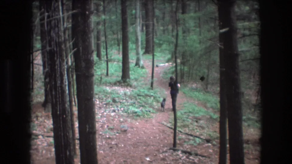 1975 Baxter, Maine home-movie frame with a woman and small dog moving away on a curved dirt path through dense green woods.