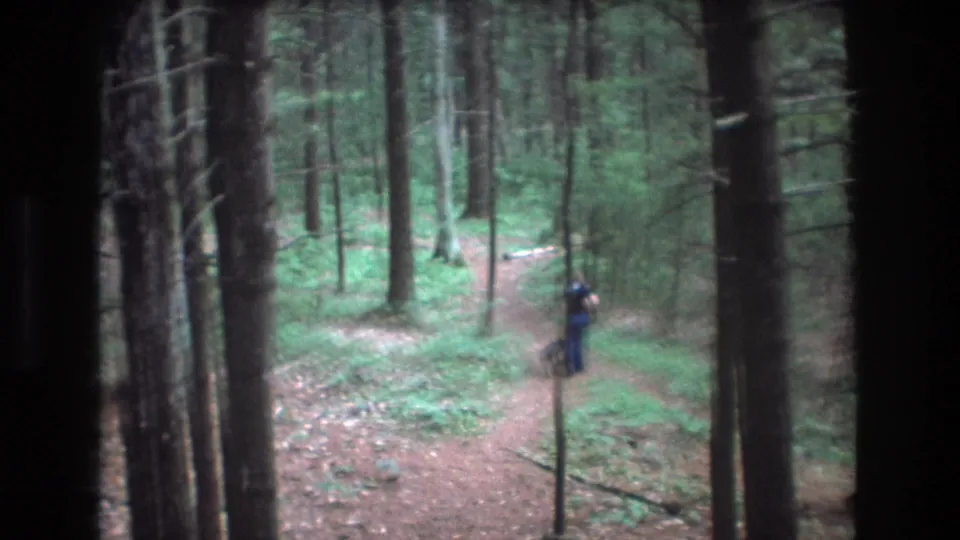 1975 Baxter, Maine 8mm frame showing a distant woman and small dog on a narrow trail, partially obscured by tree trunks and dark frame edges.