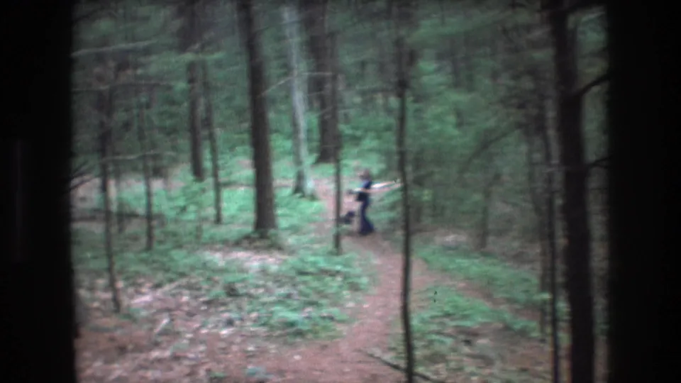 A distant walker and small dog appear on a wooded trail in the Baxter, Maine 1975 8mm footage, partly obscured by trees and motion blur.