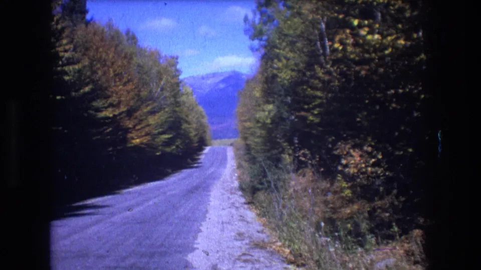 A forward-facing travel shot shows a paved rural road bordered by dense trees, with distant mountains and bright sky visible in the Baxter, Maine landscape.