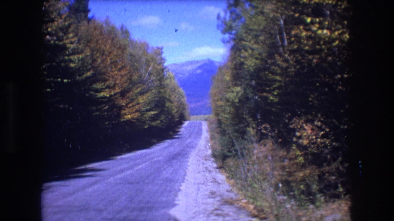 1975 8mm home-movie frame from Baxter, Maine showing a quiet paved road bordered by dense fall trees, with blue mountains visible beyond the road.