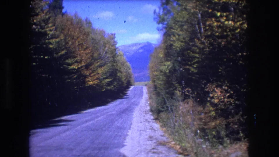 1975 8mm home-movie frame from Baxter, Maine showing a quiet paved road bordered by dense fall trees, with blue mountains visible beyond the road.