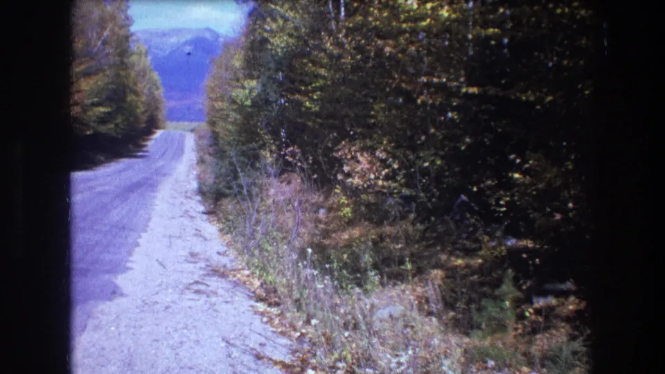 1975 8mm frame from Baxter, Maine showing the road shoulder and dense autumn trees, with the road receding toward a distant mountain view.