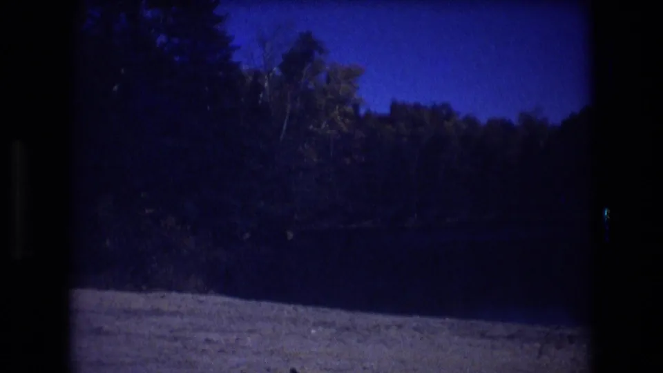 Final 1975 Baxter, Maine home-movie frame showing a dark lake or open water area with forested shoreline and hillside beneath a deep blue sky.