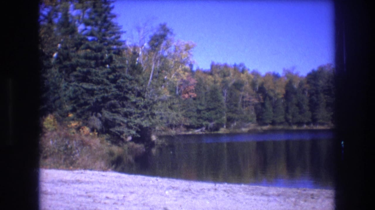 1975 Baxter, Maine 8mm frame showing a still lake, sandy foreground shoreline, evergreen trees, autumn-tinted foliage, and clear blue sky within dark film borders.